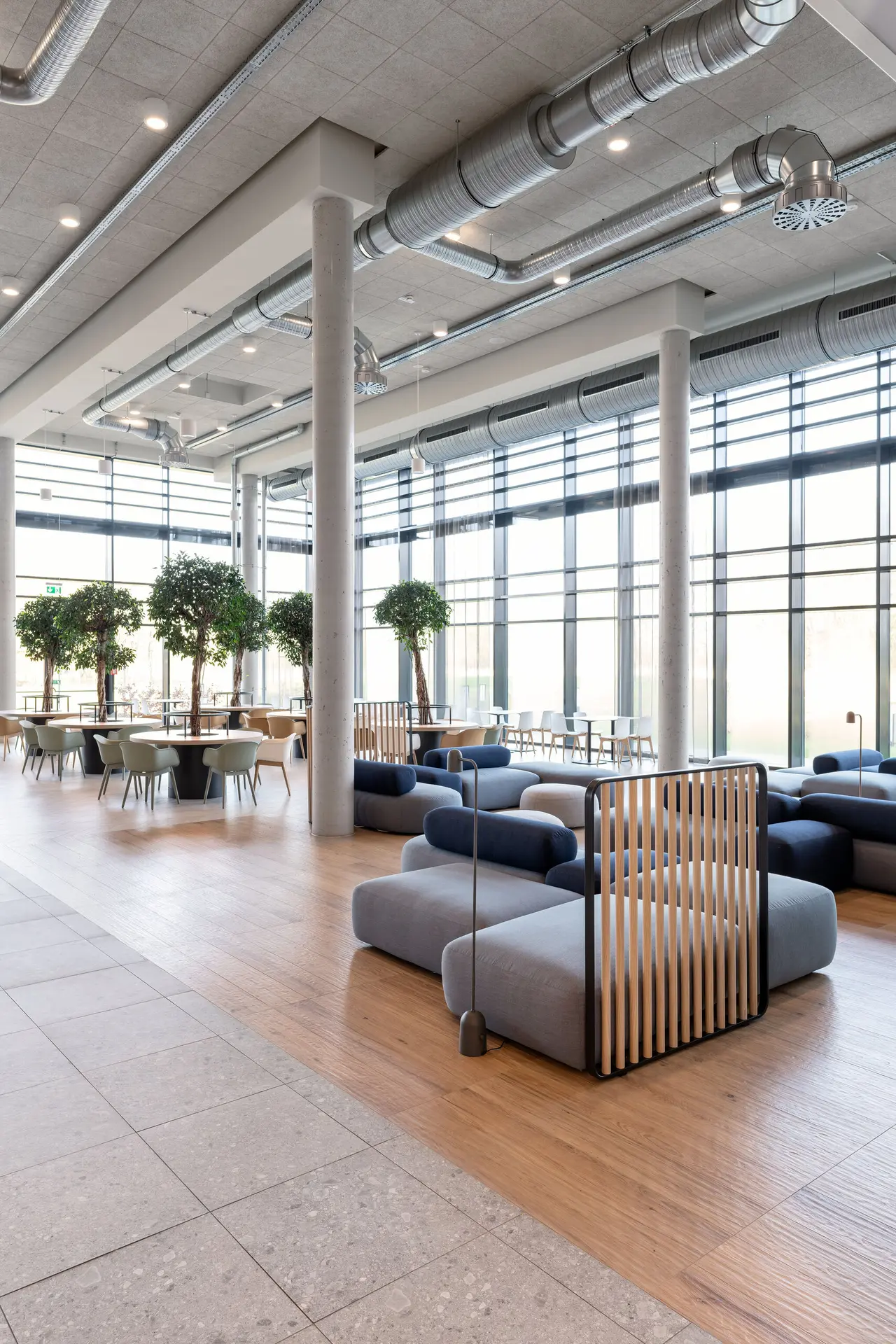 Öffentlicher Aufenthaltsbereich mit Softseating, Schalensesseln und Tischen mit Bäumen.